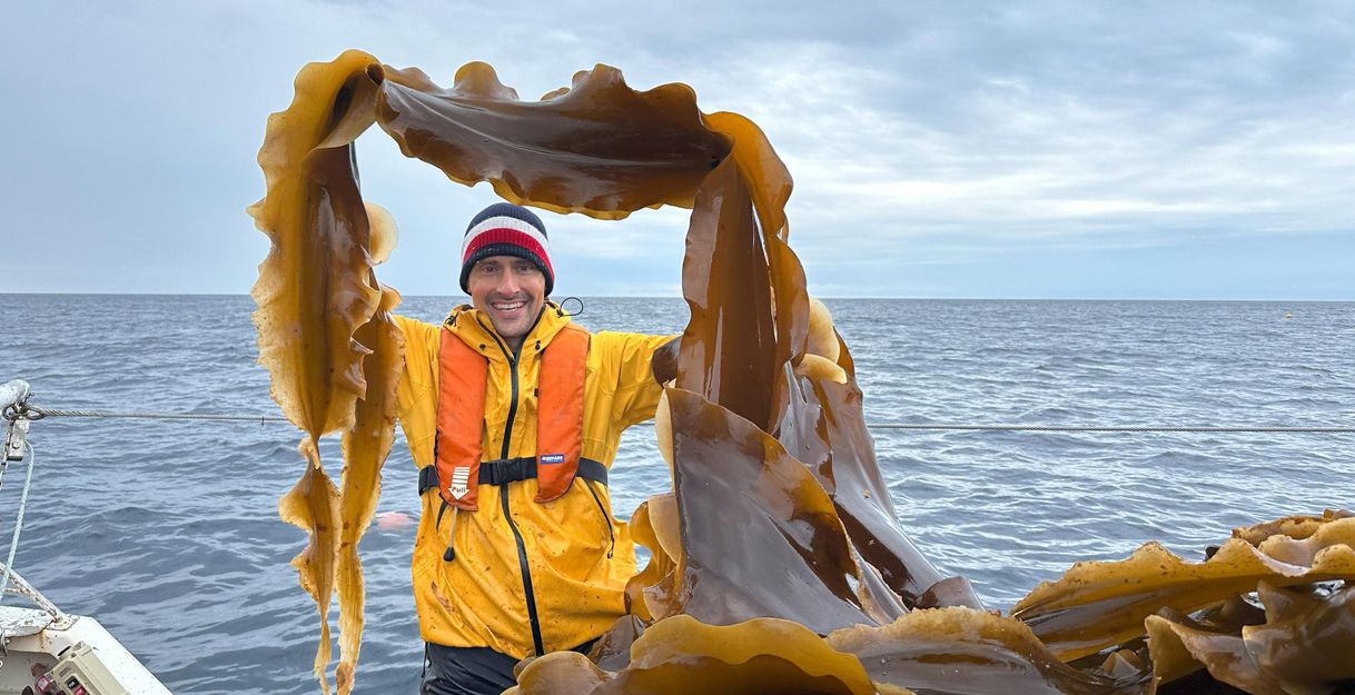 A cheerful man in a yellow rain jacket stands on a boat in the sea. He is holding seagrass in his hand and smiling under a cloudy sky.