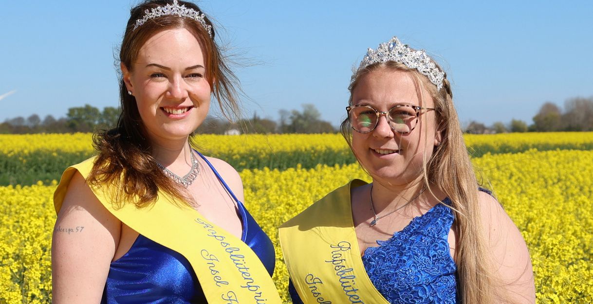 Zwei Frauen in blauen Kleidern und gelben Schärpen stehen in einem Rapsfeld.