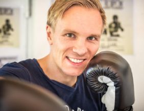 A smiling man with boxing gloves stands in a boxing studio. In the background, various boxing posters can be seen.