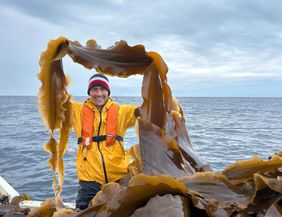 A cheerful man in a yellow rain jacket stands on a boat in the sea. He is holding seagrass in his hand and smiling under a cloudy sky.