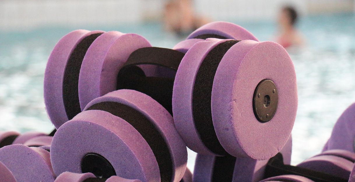 Schwimmende Hanteln in lila Farbe liegen am Beckenrand. Im Hintergrund ist ein Schwimmbad zu sehen.