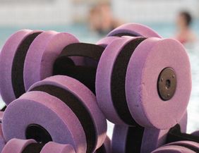Schwimmende Hanteln in lila Farbe liegen am Beckenrand. Im Hintergrund ist ein Schwimmbad zu sehen.