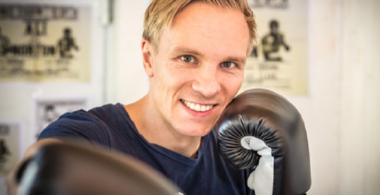 A smiling man with boxing gloves stands in a boxing studio. In the background, various boxing posters can be seen.
